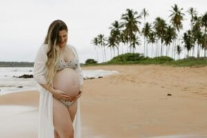 Smiling pregnant woman in bikini on sandy beach with palm trees in background.